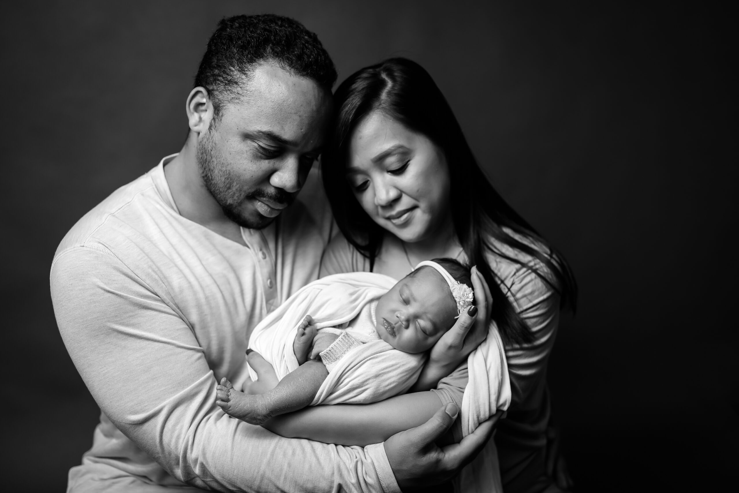 Emotional black and white portrait of a mother and father tenderly holding their newborn baby girl, wrapped in a soft blanket and wearing a floral headband. Photographed in Los Angeles by Kendra Greenberg Photography.