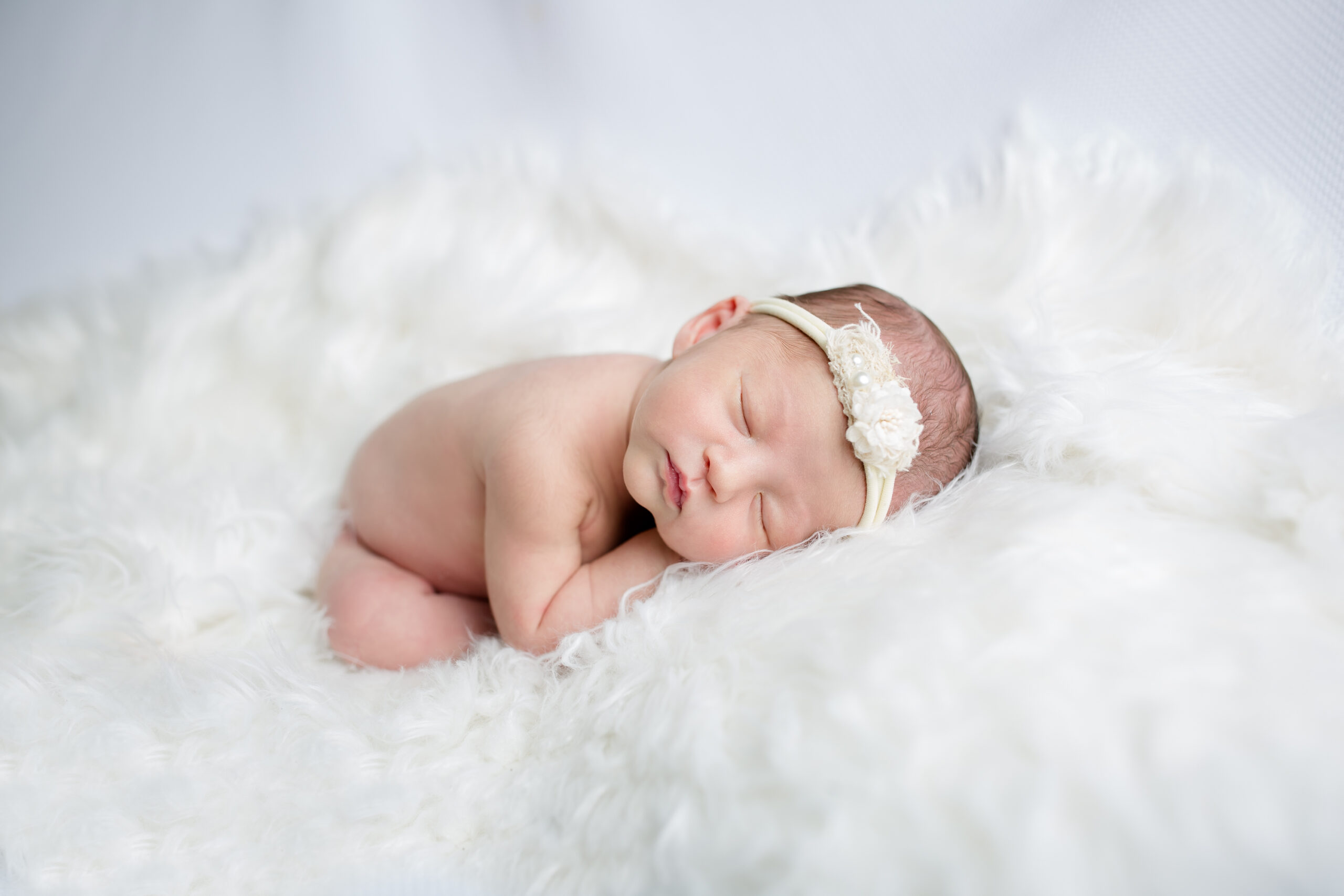 Newborn baby girl sleeping on a soft white blanket, wearing a delicate white headband, photographed in a bright Los Angeles studio by Kendra Greenberg Photography.