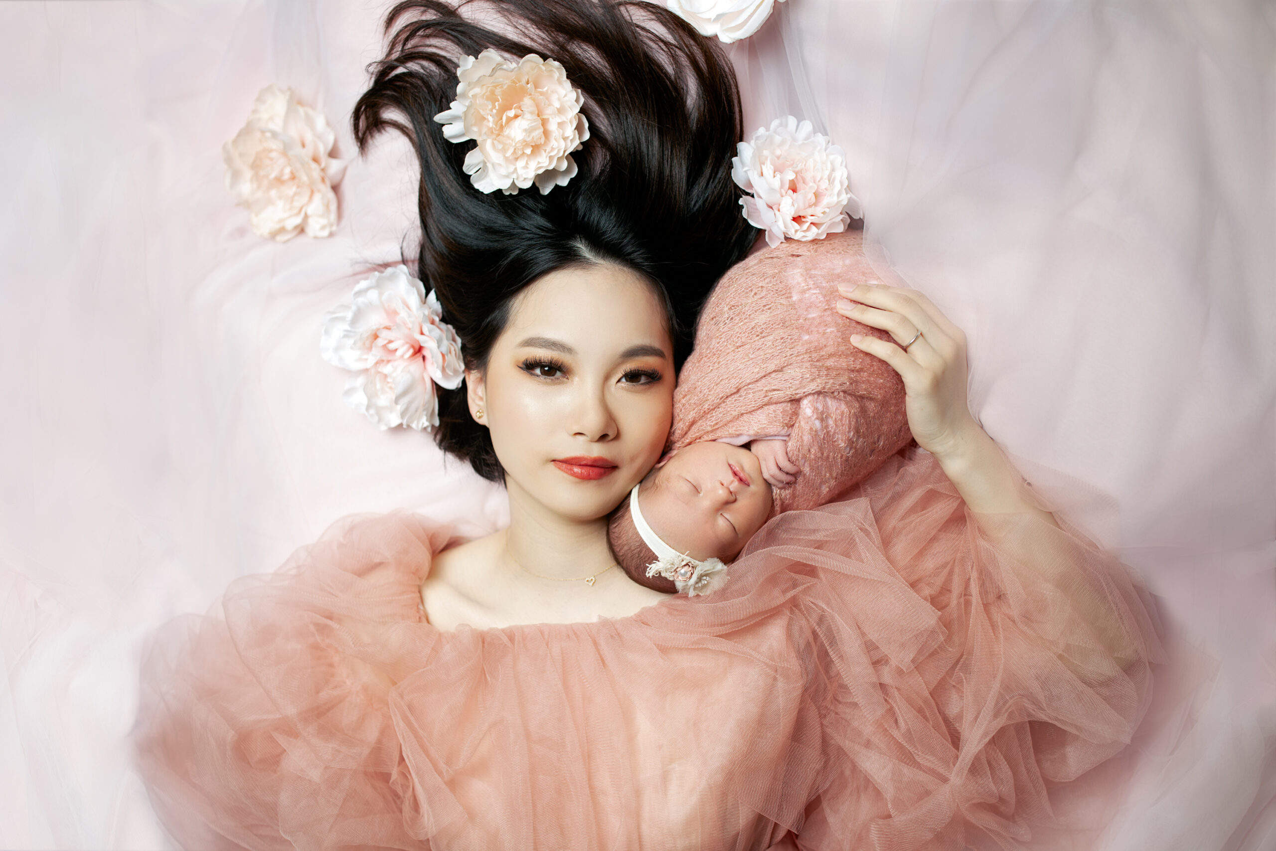 legant mother in a soft pink tulle gown lies beside her newborn baby, who is swaddled in matching tones and surrounded by delicate flowers. Photographed in Los Angeles by Kendra Greenberg Photography.
