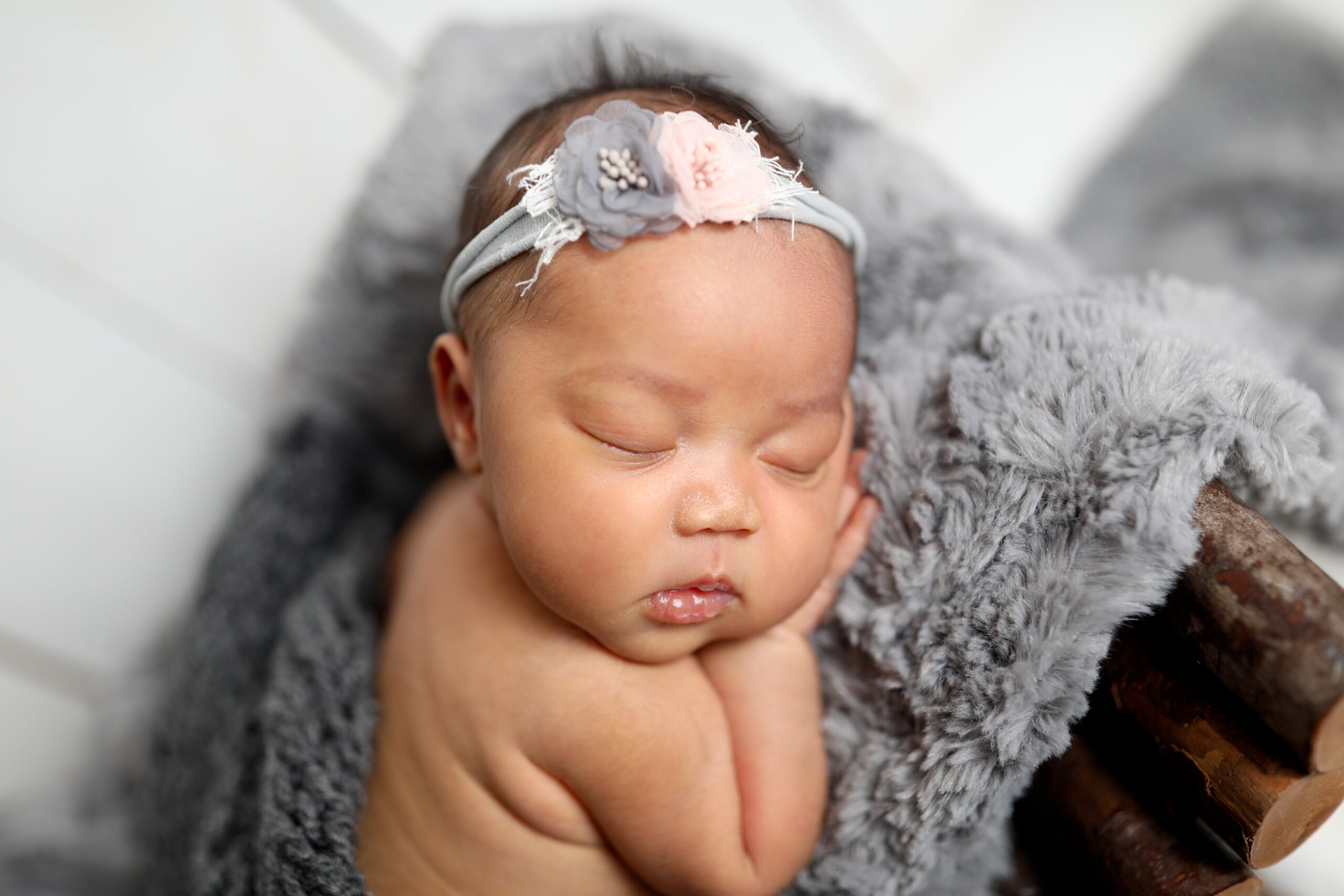 Close-up of a sleeping newborn baby with a soft gray and pink floral headband, resting on a fluffy gray blanket. Photographed in Los Angeles by Kendra Greenberg Photography.