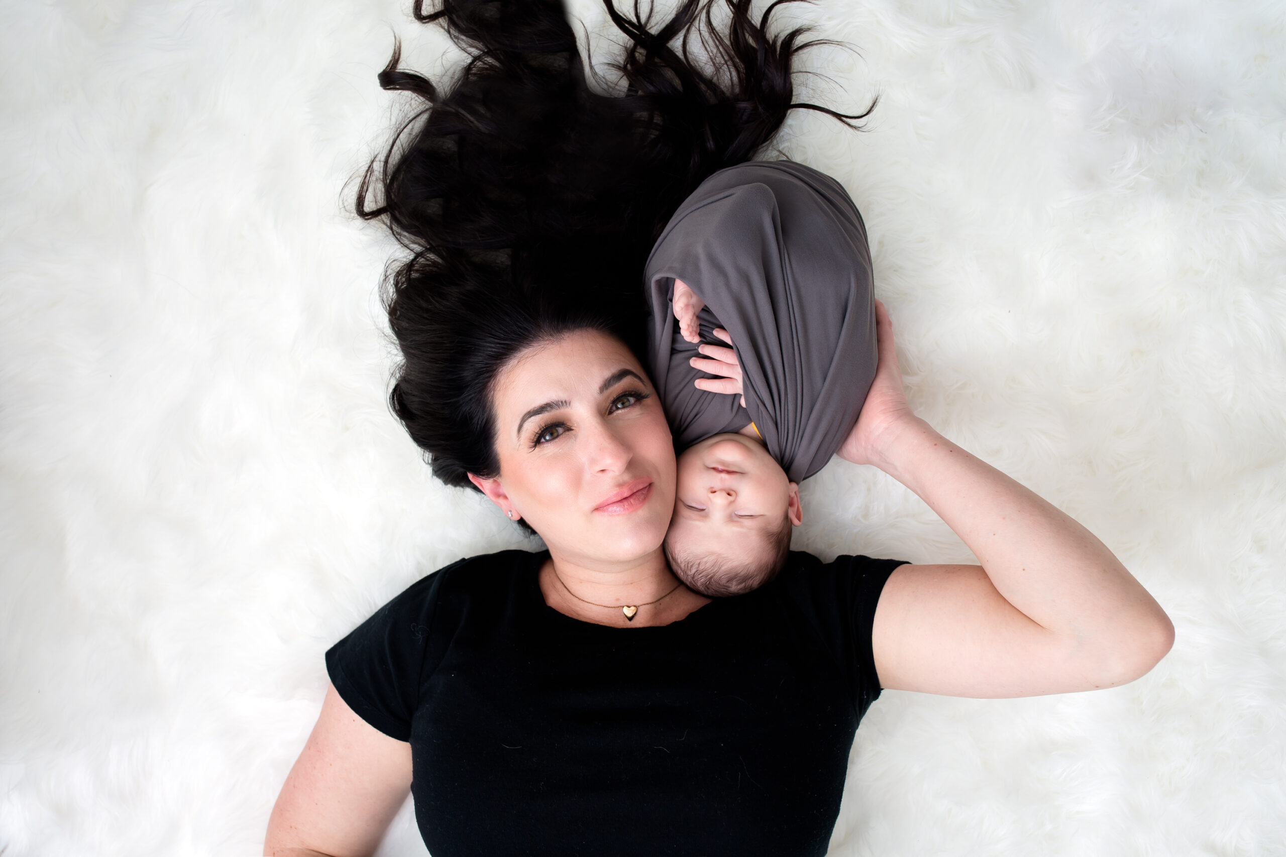 Mother lying on a soft white fur rug with her newborn baby swaddled in gray, their faces gently touching as they gaze peacefully. Photographed in Los Angeles by Kendra Greenberg Photography.