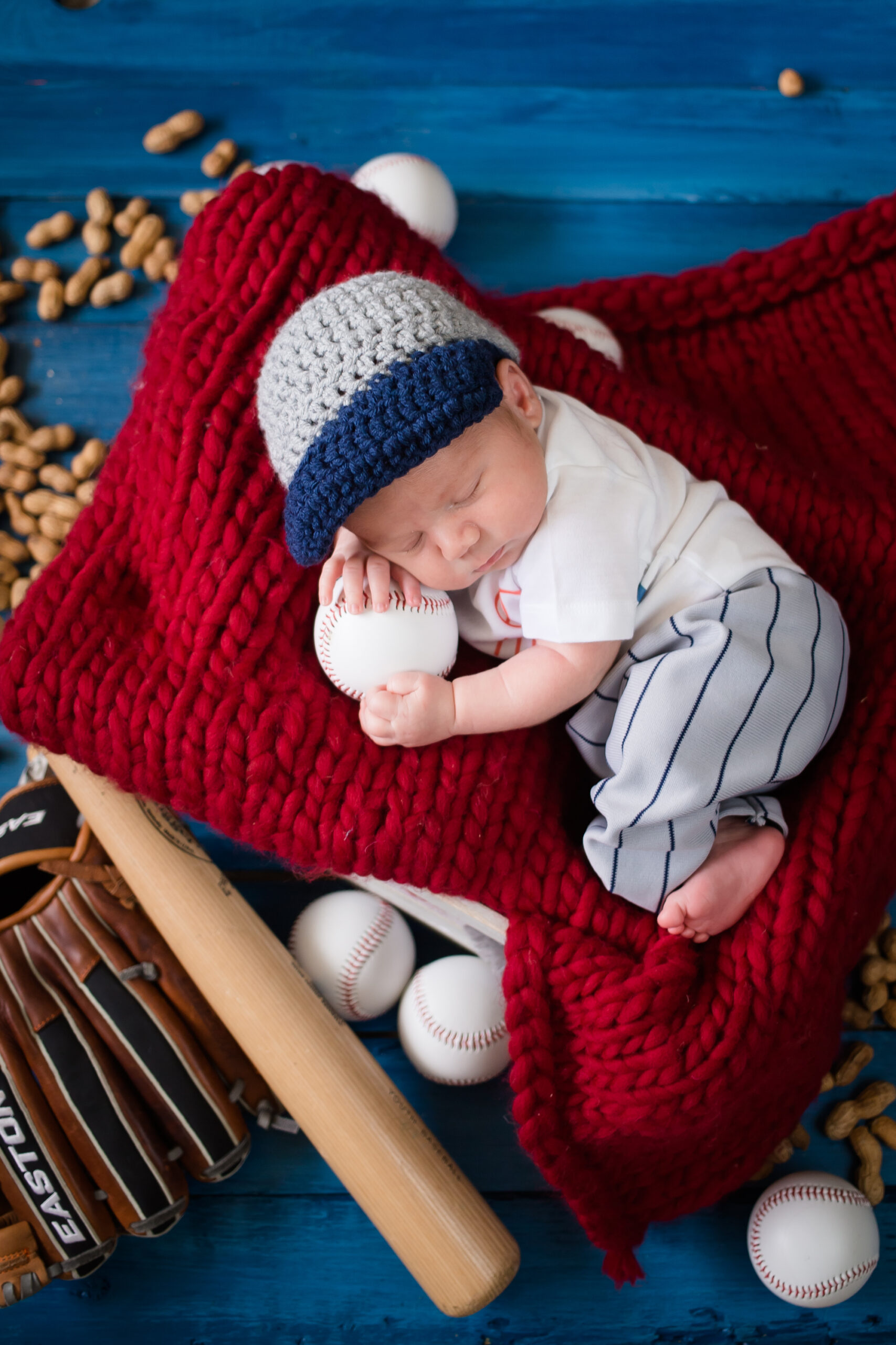 Newborn baby dressed in baseball-themed outfit, cuddling a baseball while sleeping on a red knit blanket surrounded by peanuts, a bat, glove, and baseballs. Photographed in Los Angeles by Kendra Greenberg Photography.