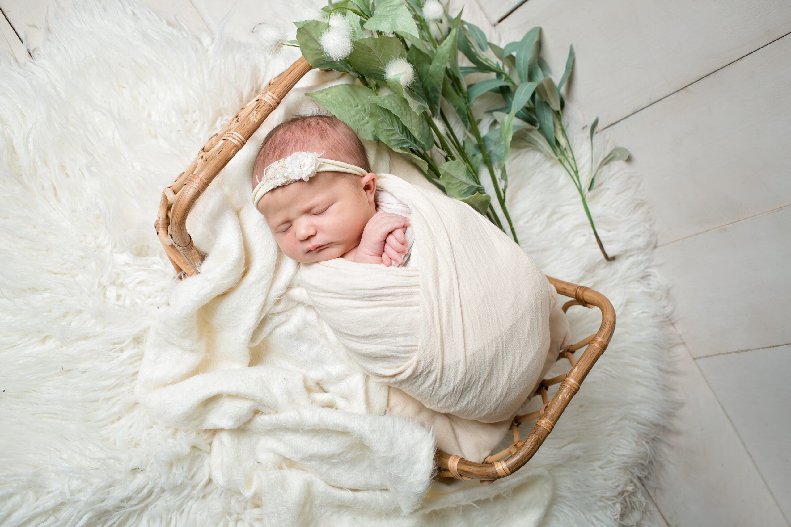 Newborn baby peacefully swaddled in a cream wrap, sleeping in a small wicker basket with white blankets and greenery, photographed in a bright, minimal Los Angeles studio.